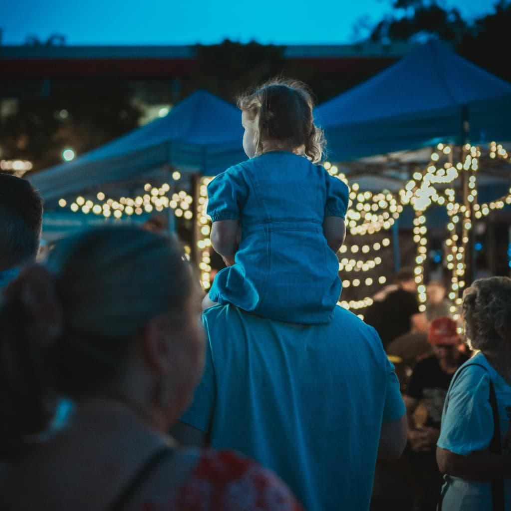Child on an adult's shoulders at a twilight market, fairy lights and stalls ahead