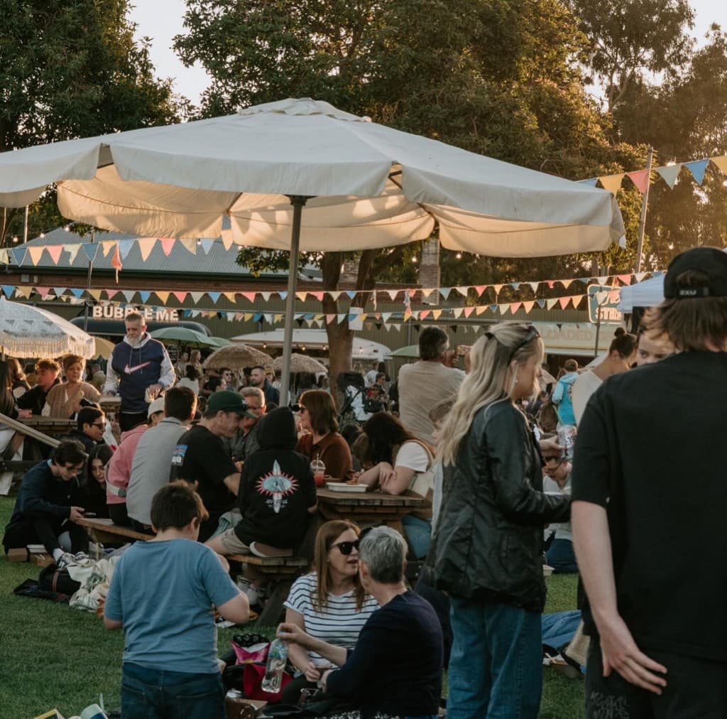 Crowd at an outdoor market on the grass at golden hour, picnic tables and bunting